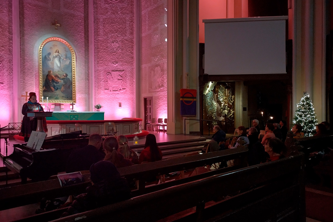 Kirkon alttari on valaistu punaisen sävyin, edessä puhuu ihminen ja ihmisiä istuu kirkon penkeissä.