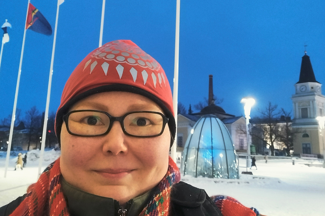 Nainen hymyilee ja katsoo kameraab. Taustalla lipputankoja ja lippu sekä kirkko.