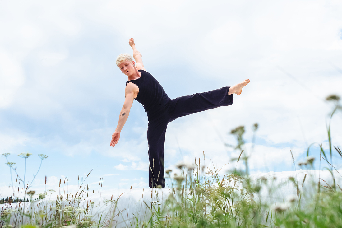 Mies mustissa vaatteissa tanssii kesäisellä nurmikolla ja taustalla sininen taivas.