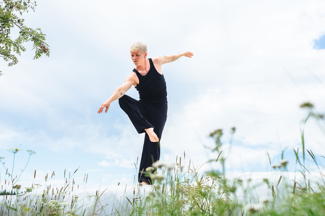 Mies mustissa vaatteissa tanssii kesäisellä nurmikolla ja taustalla sininen taivas.