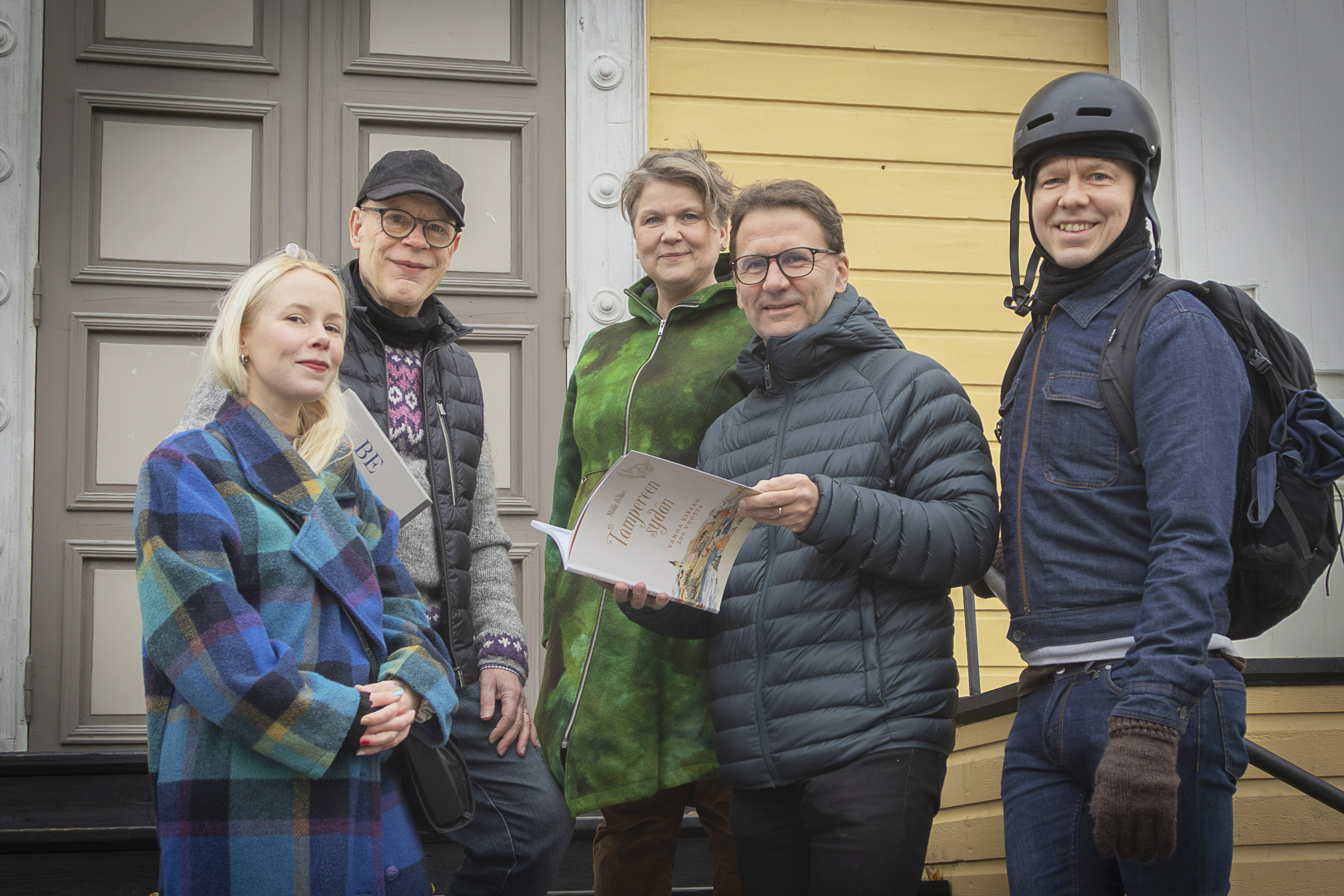 Kirkon portailla seisoo viisi ihmistä, joista yksi tarkastelee kirjaa ja yksi on pukeutunut pyöräilyasuun ja kypärään.