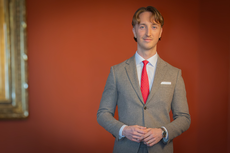 Mies puku päällä seisoo punaisen seinän edessä ja hymyilee kameralle.