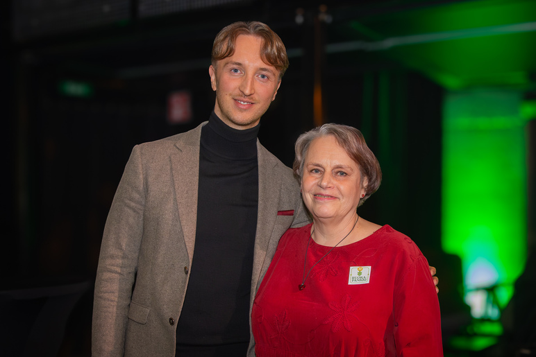 Mies ja nainen seisovat tummahkossa tilassa ja mies pitää kättään naisen olkapäällä.