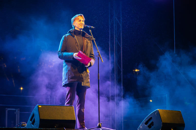 Mies puhumassa lavalla illalla. Lavalla siniset valot ja hiukan sumua.