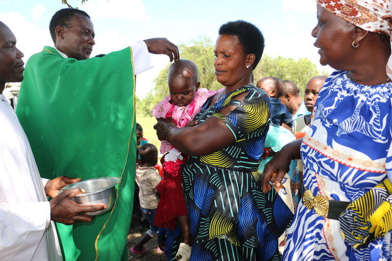 Kaapuun pukeutunut mies valaa vettä pienen lapsen päähän, ja toinen mies pitelee vieressä astiaa. Lapsi on värikkääseen mekkoon pukeutuneen naisen sylissä, ja toinen nainen puhuu vieressä. Taustalla on lapsia.