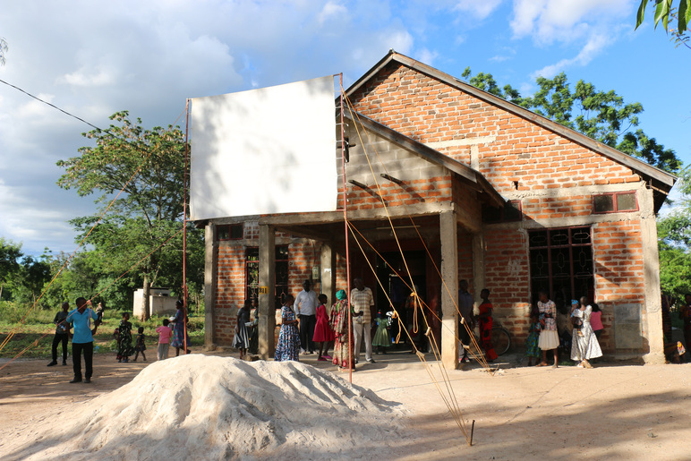 Punasavitiilisen rakennuksen edessä on kasa hiekkaa ja teline, johon on kiinnitetty suuri tyhjä taulu. Seinien vierillä on ihmisiä.
