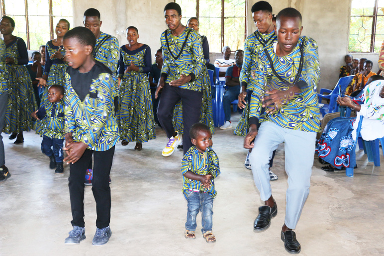 Monen ikäisiä ihmisiä tanssii samasta kankaasta tehdyissä paidoissa tai mekoissa sisällä rakennuksessa. Kaksi tanssijoista on pikkulapsia. Takana istui ihmisiä muovituoleissa.