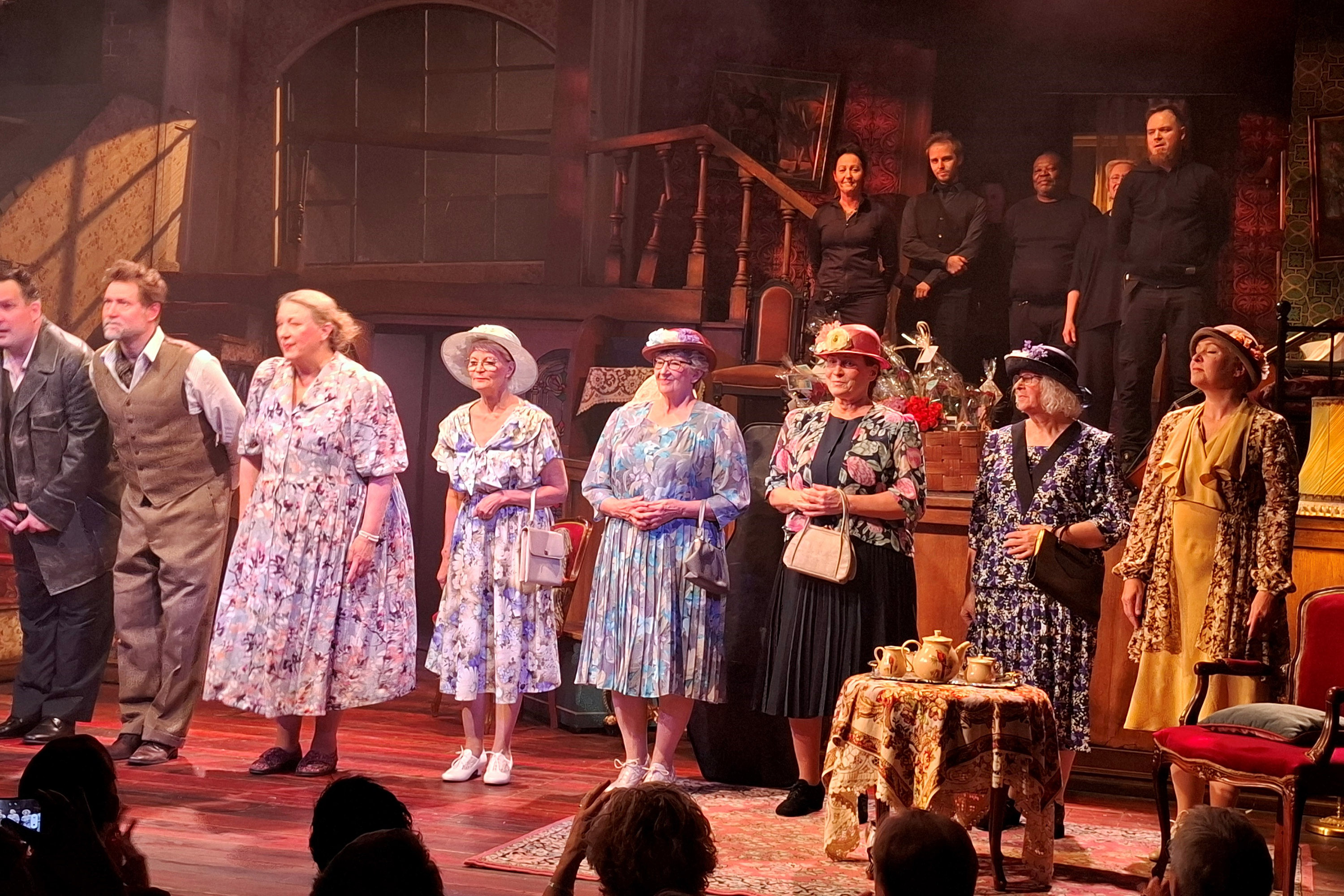 Kuusi naista ja kaksi miestä näkyy näyttämöllä rivissä. Naisilla on mekot ja hatut, ja heistä neljällä myös käsilaukku. Etualalla sivussa on pieni pyöreä pöytä, jonka päällä on kahvikuppeja.