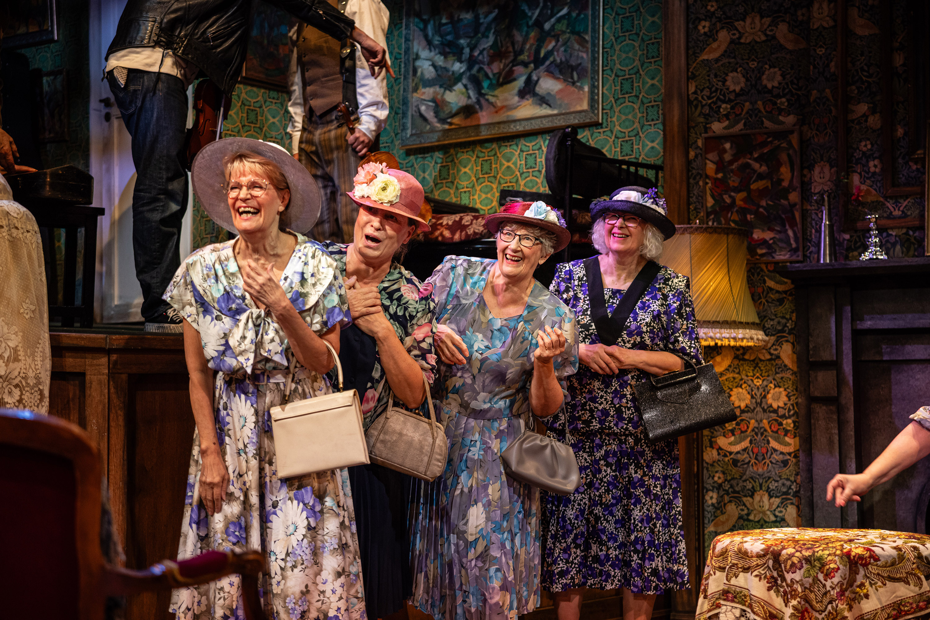 Neljä nauravaa naista, joilla on mekot, hatut ja käsilaukut, seisoo pienessä tilassa, jonka seinät on peitetty antiikkisilla tapeteilla.