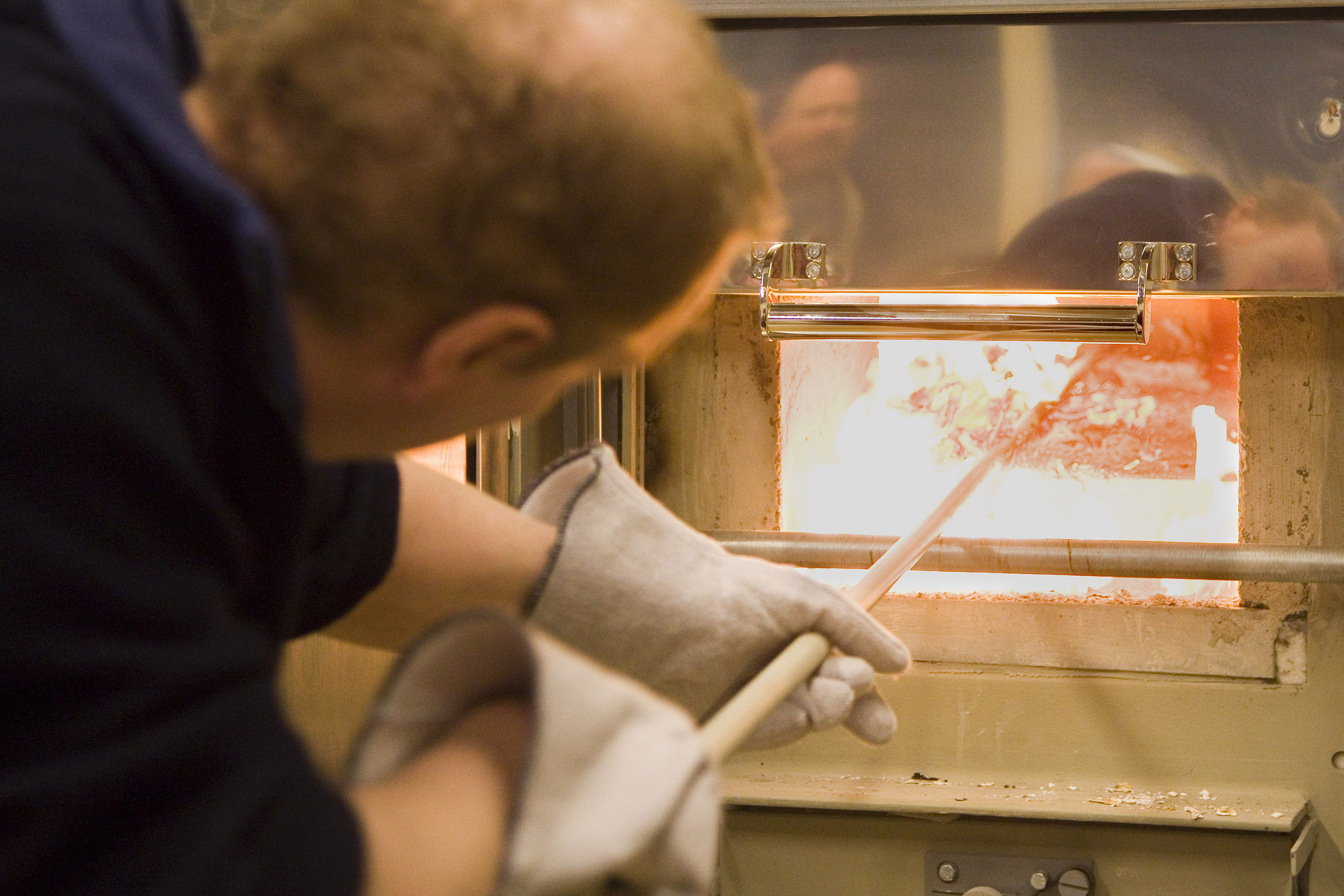 Lähikuva uunin pesästä, jossa palaa tuli. Joku käsittelee tulta isolla tuhkakaapimella kintaat käsissään.