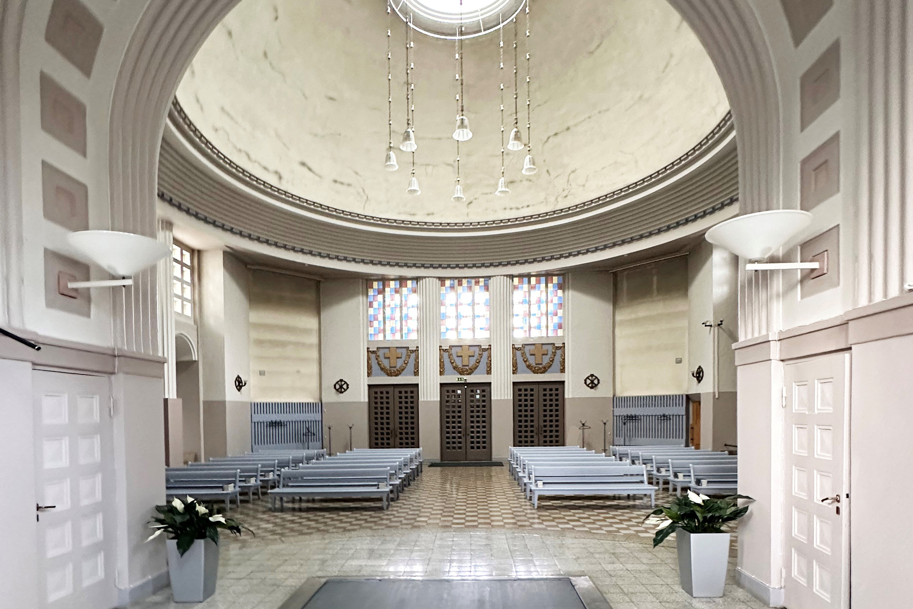 Rakennuksen sisäkuva kohdasta, jossa on kaareva syvennys, sinne vievät portaat ja syvennyksessä lattialla seisova risti sekä kaksi pitkää kynttilänjalkaa kynttilöineen.