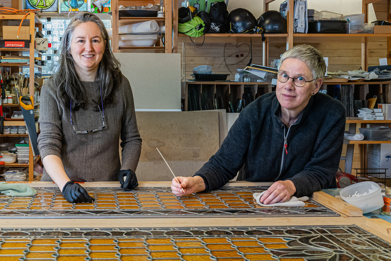 Kaksi henkilöä seisoo edessään työpöytä, jonka täyttää lasipaloista ja niiden ristikkomaisista metallikehyksistä koostuva rakenne. Vasemmanpuoleisella on käsineet, oikeanpuoleisella toisessa kädessä puikko ja toisessa liina.