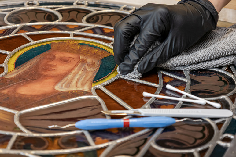 Käsineellä suojattu käsi puhdistaa liinalla taideteosta, joka koostuu värikkäistä lasipaloista ja niitä ympäröivistä metallikehyksistä. Kuvassa on myös työn yksityiskohta, jossa on henkilöhahmo päässään kruunu.