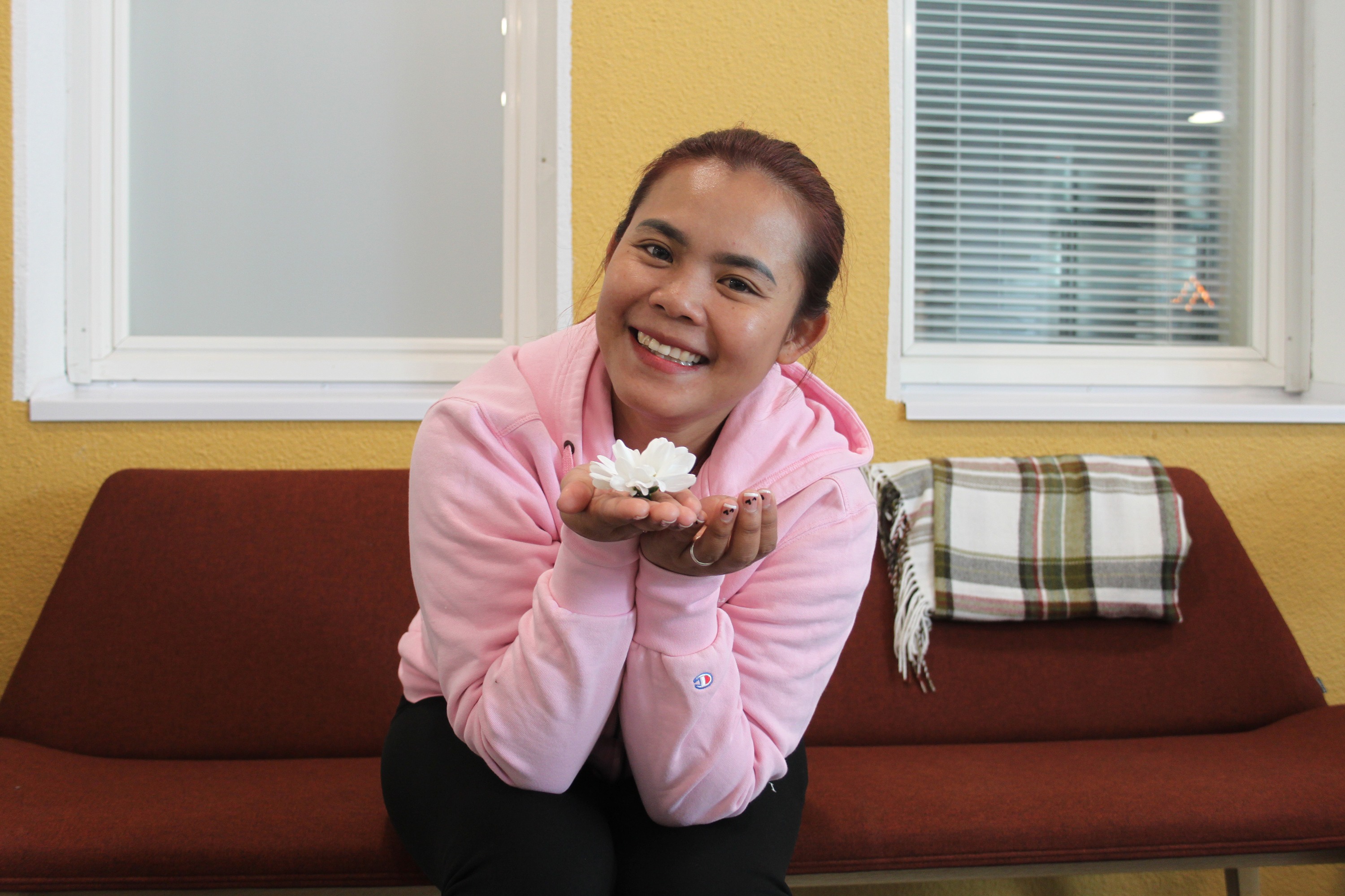 Hymyilevä sohvalla ikkunoiden edessä istuva nainen pitää käsissään kukkaa ja ojentaa sitä eteenpäin.