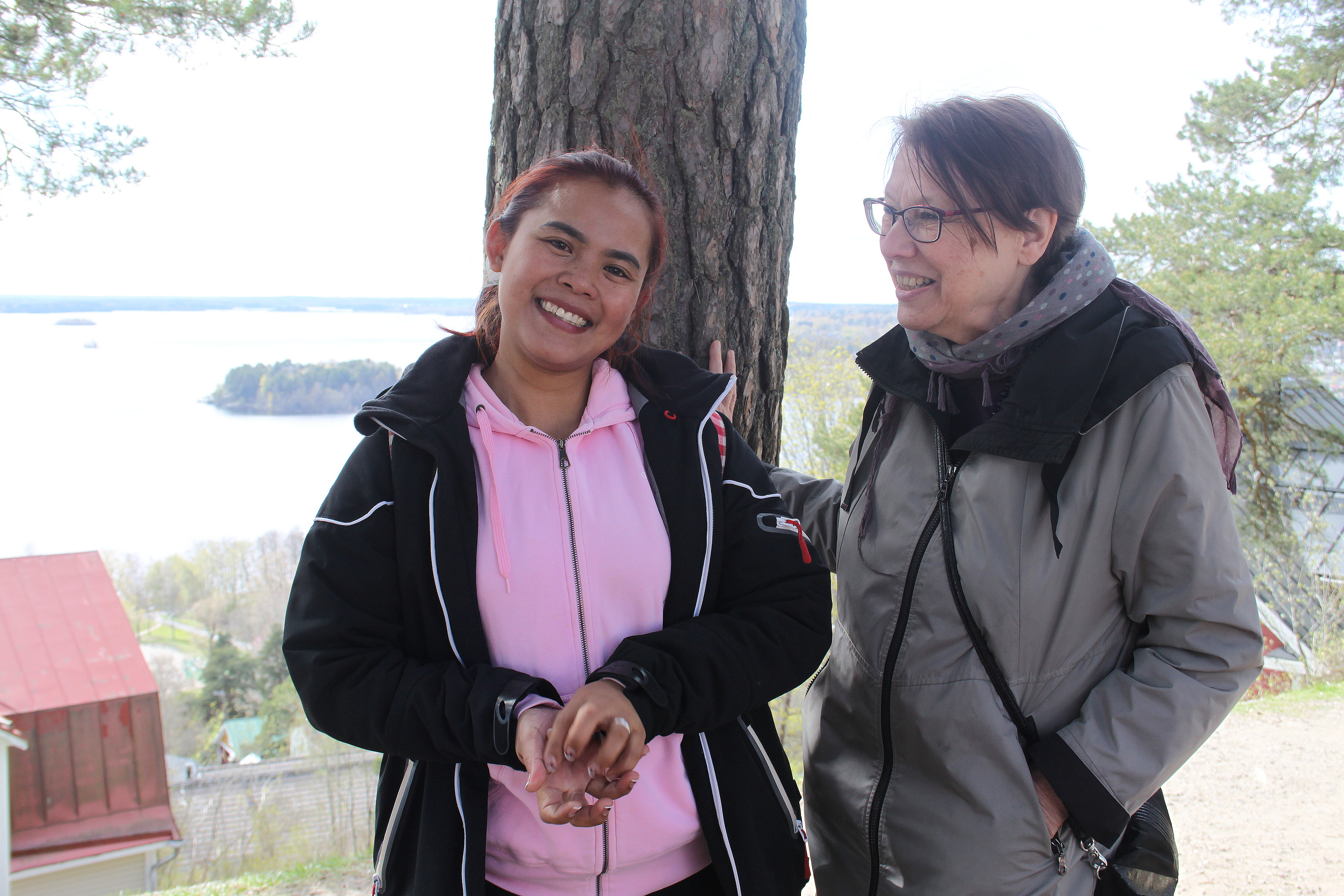 Kaksi naista, joilla on takit päällä, seisoo puun luona avoimen järvimaiseman edessä.