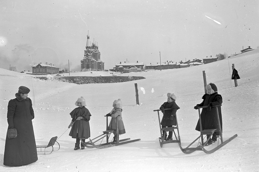 Mustavalkoinen kuva, jossa lapsia potkukelkoilla lumisen rinteen edustalla. Aikuinen seisoo vieressä pitkässä talvitakissa. Taustalla kirkko ja puutaloja.