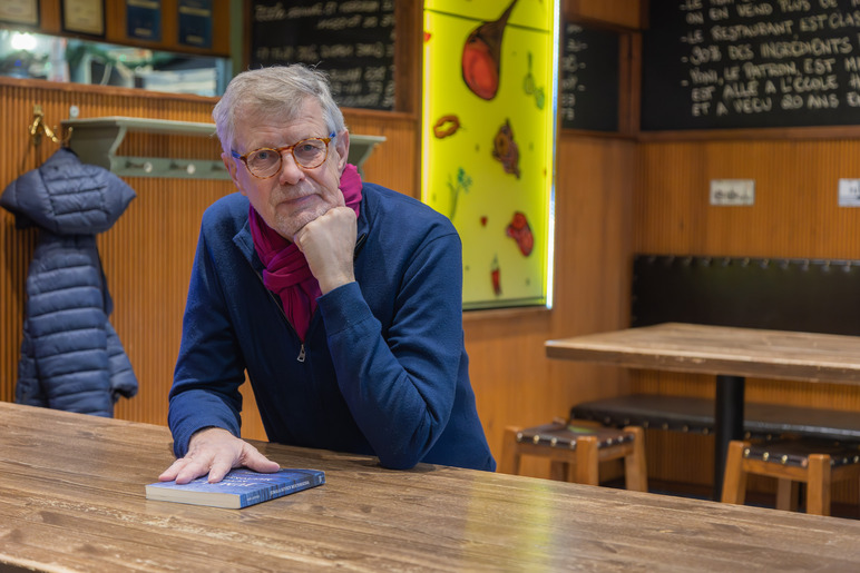 Mies istuu käsi leuan alla puisen pöydän ääressä tilassa, jossa on puisia sisustuselementtejä. Toisen kätensä alla hänellä on kirja. Taustalla on naulakko, jossa roikkuu takki.