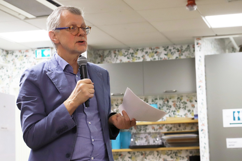 Pikkutakkiin pukeutunut seisova mies puhuu mikrofoniin ja pitelee toisessa kädessään nippua papereita.