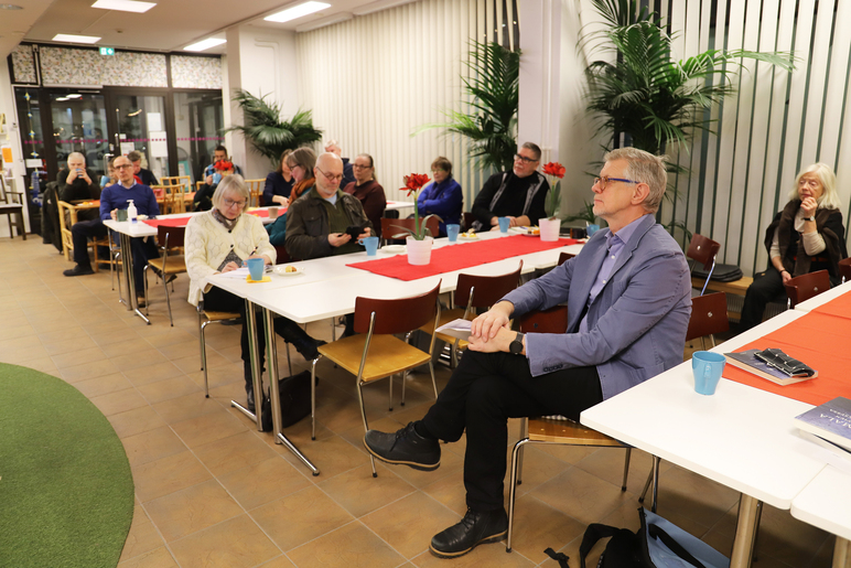 Joukko ihmisiä istuu pöytien ääressä. Etummaisena on pukuun pukeutunut mies, jolla on kädet ristissä polvien edessä.