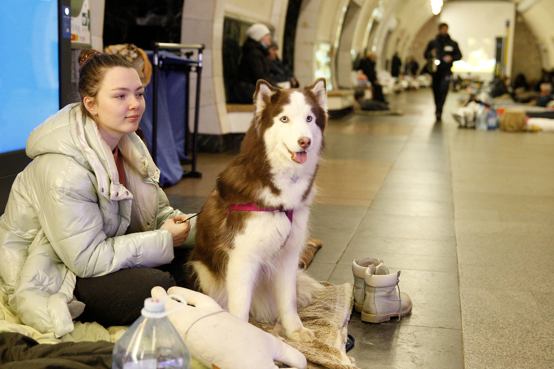 Nainen istuu metroaseman käytävällä koiransa kanssa.