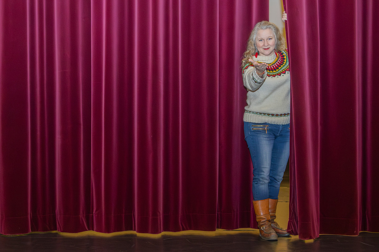 Nainen kurkistaa teatterin lavalla verhojen välistä ja kutsuu käsimerkein luokseen.