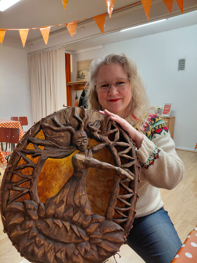 Nainen pitää isoa itse tekemää rumpua sylissään ja hymyilee.