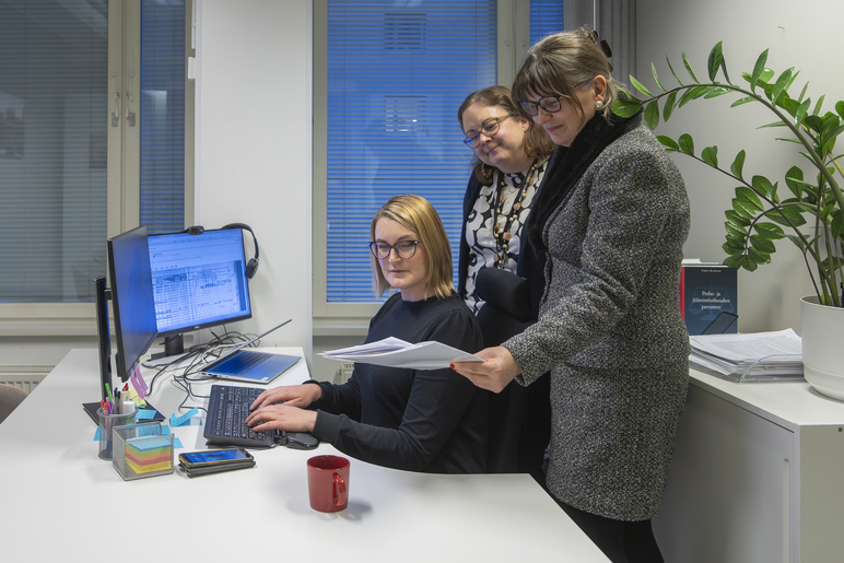 Pöydän ääressä ikkunan edessä on kolme naista, joista kaksi seisoo. Heistä oikeanpuoleinen pitää käsissään papereita, joita tietokoneen ääressä istuva vilkaisee samalla kun kädet ovat koneen näppäimistöllä.