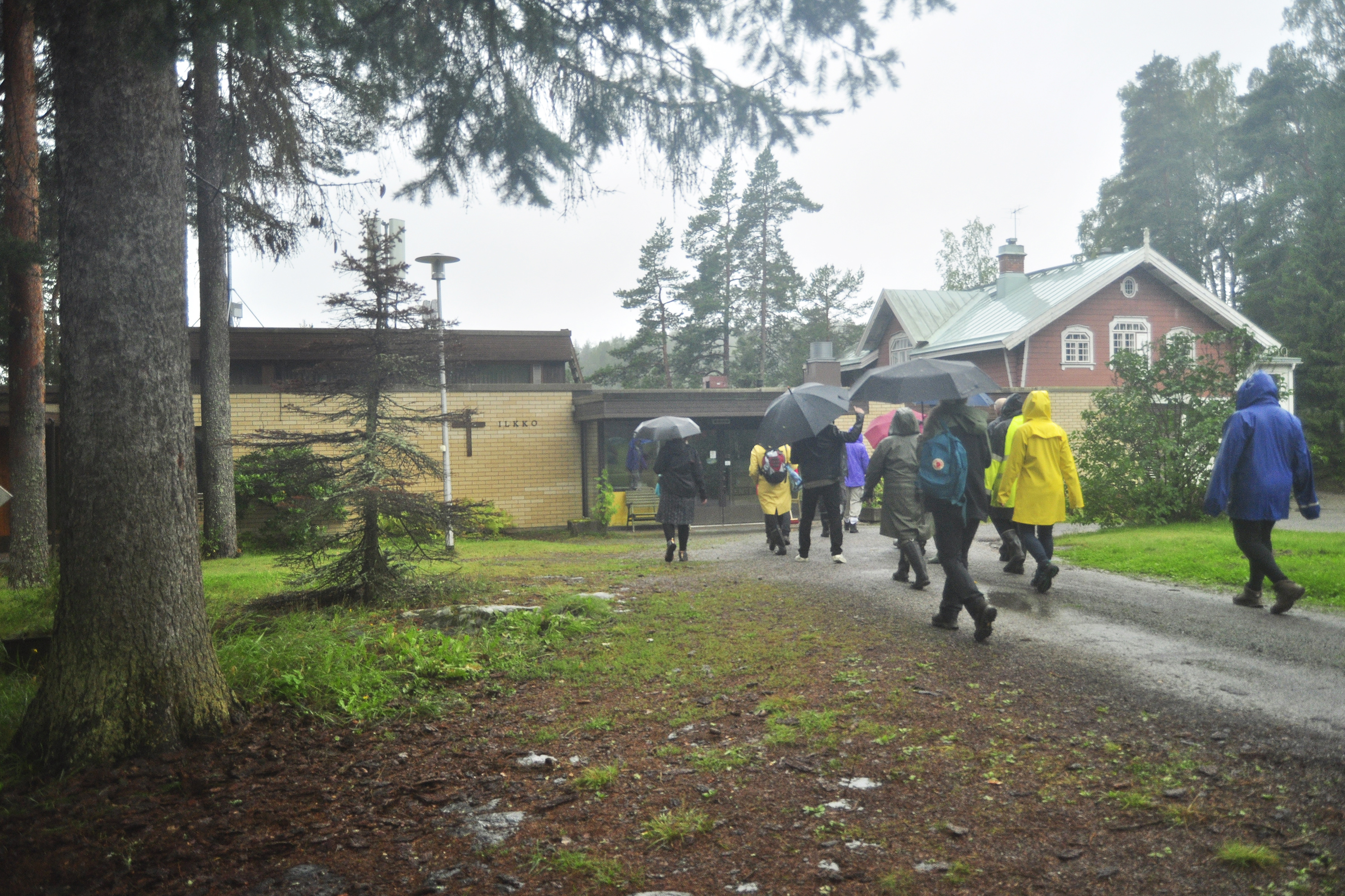 Joukko ihmisiä kävelee sateessa kohti rakennuksia, joista vasemmanpuoleinen on uudempi tiilirakennus ja oikeanpuoleinen vanhempi huvilamainen rakennus.