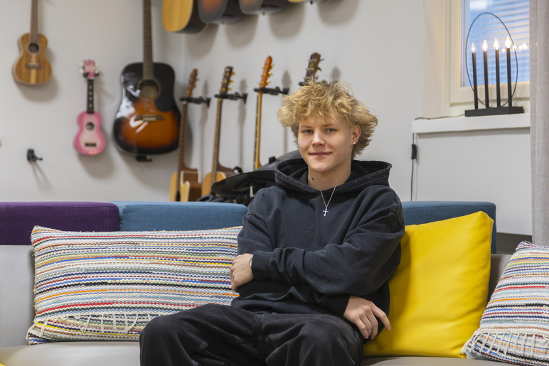 Nuori mies istuu sohvalla, jolla on isoja tyynyjä. Seinällä on monia kielisoittimia, ja ikkunalla kynttiläkruunu.