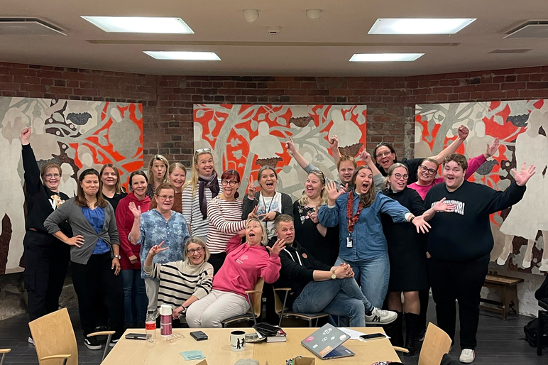 Iso joukko ihmisiä pöydän edessä seisoo tai istuu sisällä tilassa, jonka seinillä on isoja taideteoksia. Heistä osa nauraa tai ilmeilee ja heiluttaa käsiään.