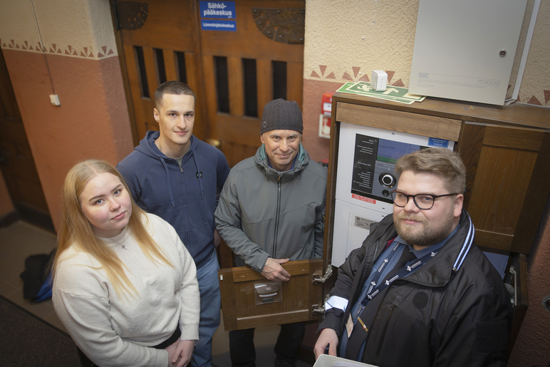 Neljä ihmistä seisoo historiallisen rakennuksen sisätilassa pystymuotoisen laitteen äärellä. Laitteen alareunassa on luukku, jonka ovi on auki.