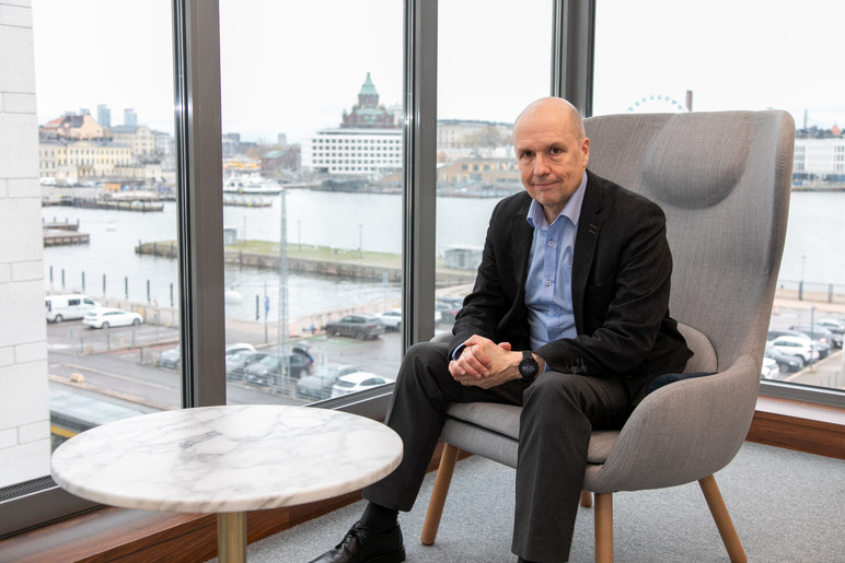 Mies istuu avaran ikkunan edessä pyöreän pienen pöydän ääressä. Ikkunasta näkyy kaupunkia, vettä ja iso laituri.