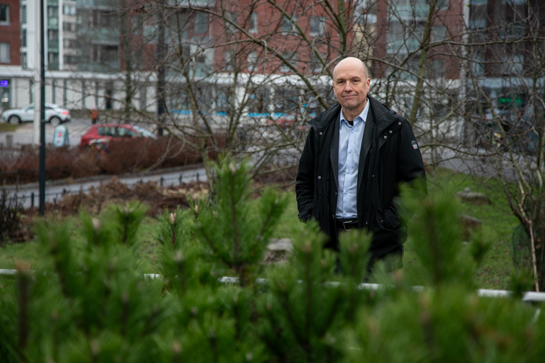 Henkilö seisoo pensaikon keskellä. Takana on kerrostalo ja autoja.