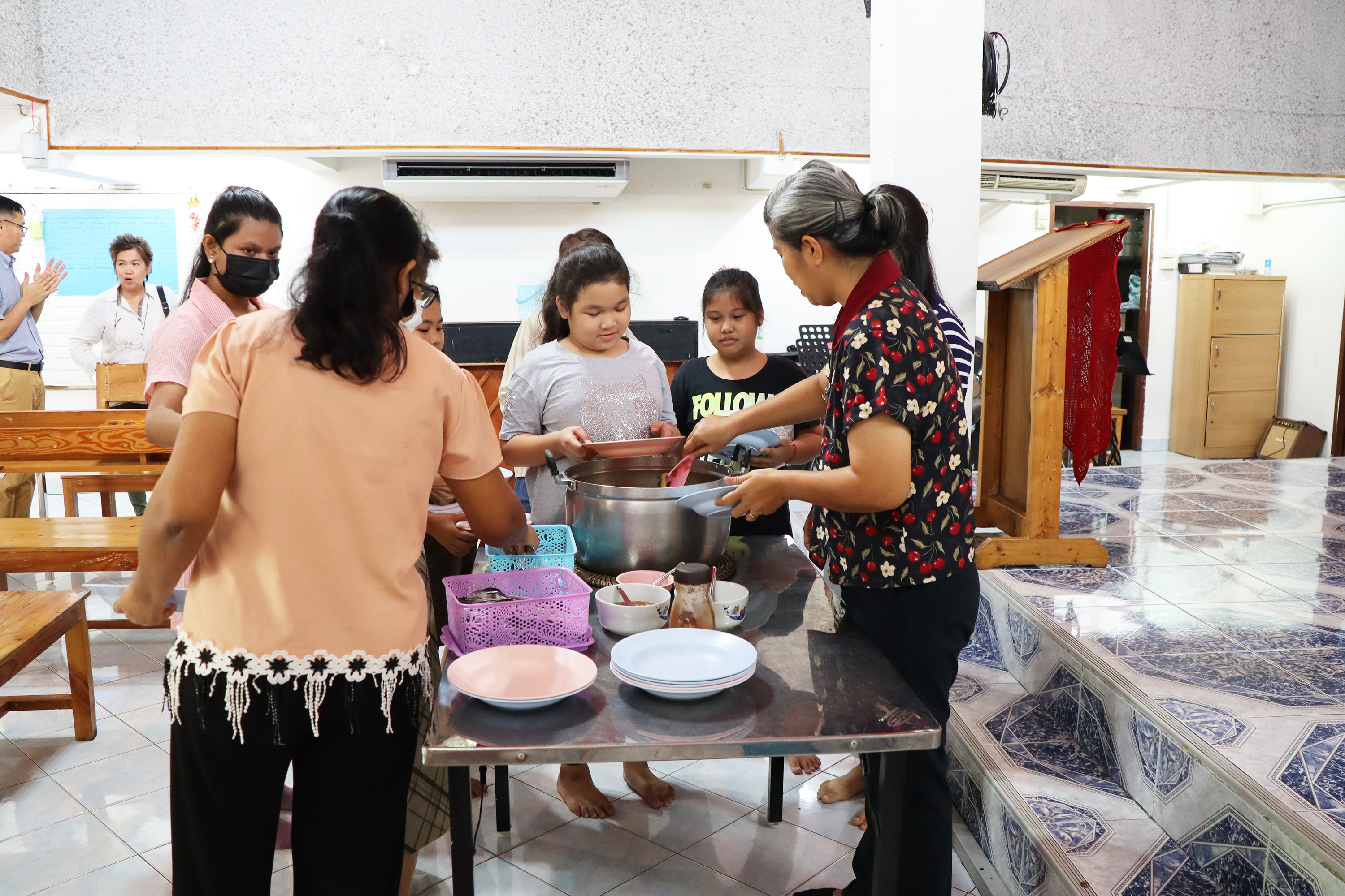 Tarjoilupöydän ääressä yksi aikuinen ottaa kauhalla ruokaa isosta kattilasta ja toinen jotain rasiasta. Pöydän takana nuorempia odottaa vuoroaan, ja taustalla keskustelee ihmisiä.