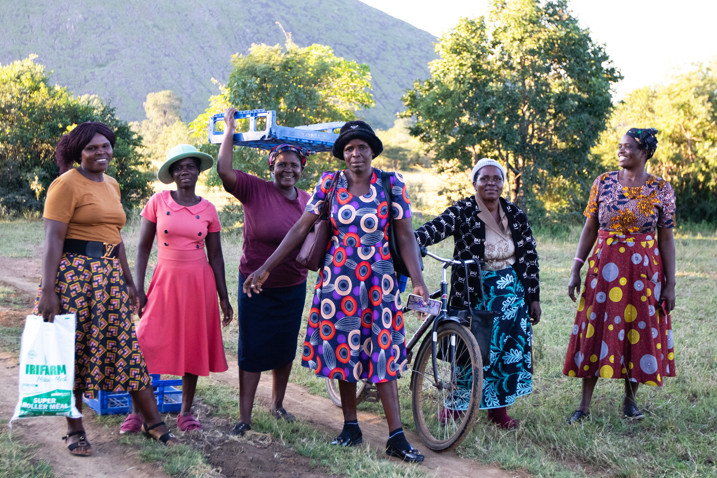 Kuusi naista rivissä ulkona. Yhdellä heistä on polkupyörä, toisella muovikassi ja kahdella muovinen kori. Kuvan keskellä kulkee polku.