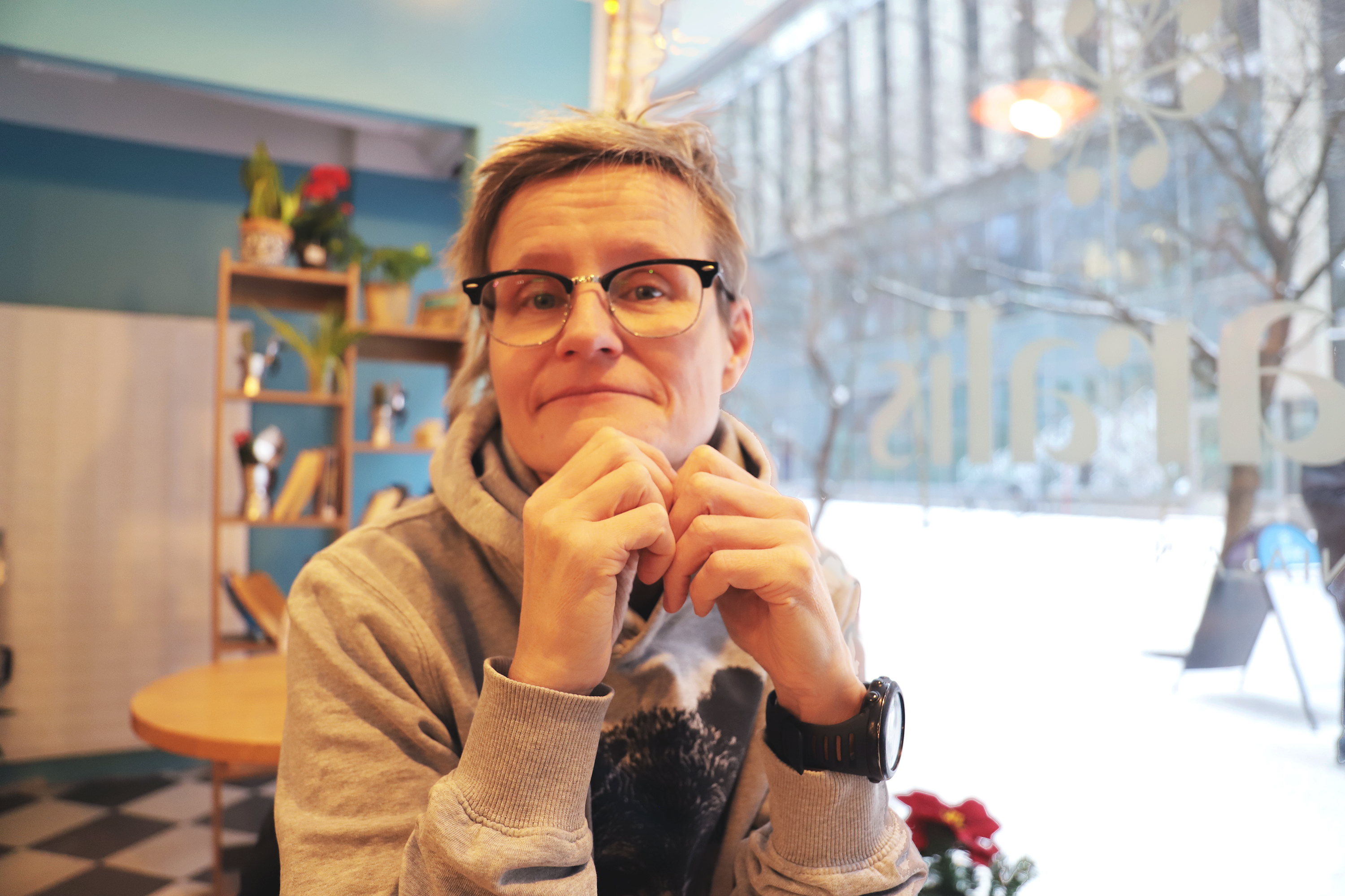 Nainen istuu kahvilassa vinosti ikkunan ääressä kädet leukojensa alla.