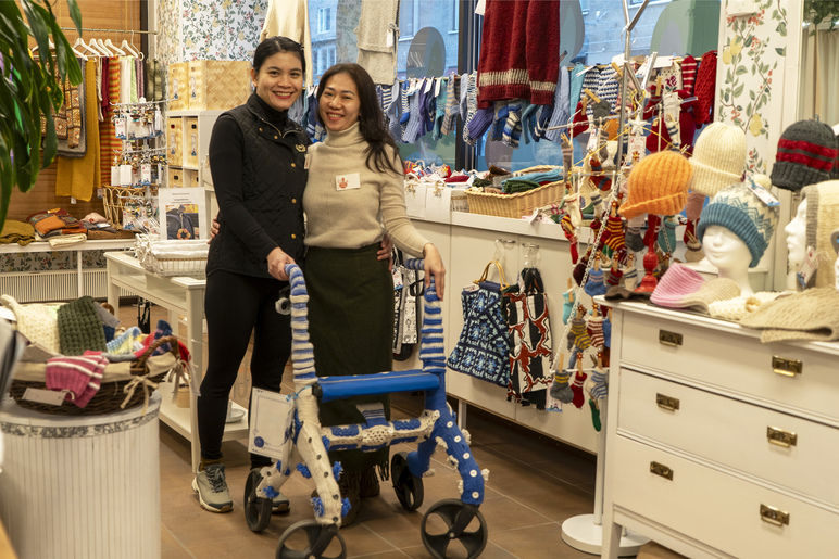 Kaksi hymyilevää naista koristellun rollaattorin kanssa huoneessa, joka pursuaa käsitöitä.