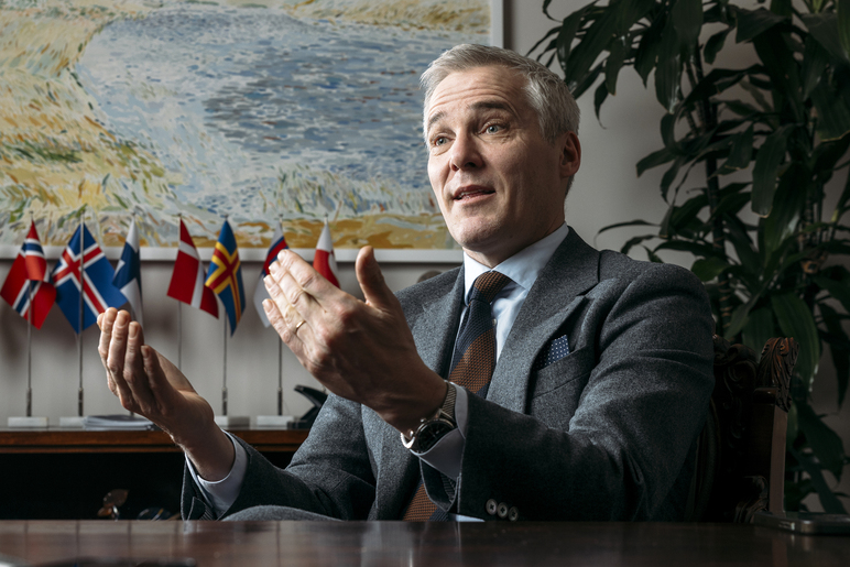 Pöydän ääressä istuva hymyilevä henkilö nostaa käsiään huoneessa, jossa on seinällä maalaus ja sen edessä tasolla maiden pöytälippuja.