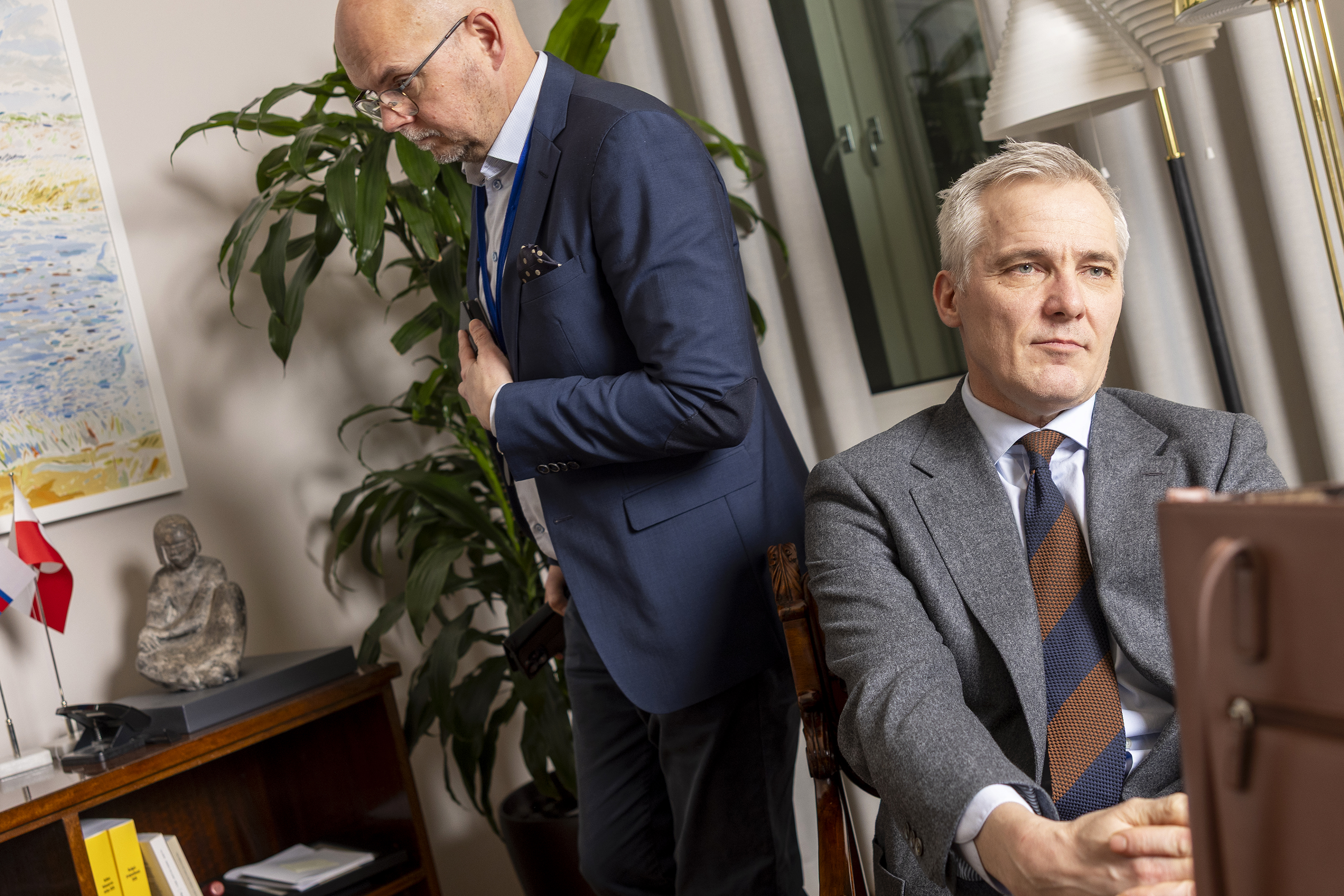 Vinoon otettu otos, jossa etualalla istuva henkilö pitää käsiään ristissä polvien ympärillä. Takana huoneessa kävelee toinen henkilö, joka pitää oikeaan kättään rinnan päällä.