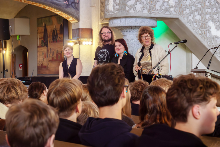 Etualalla selin istuu paljon nuoria poikia. Taempana saarnatuolin pylvään edessä seisoo neljä aikuista. Taustalla näkyy maalaus seinässä.