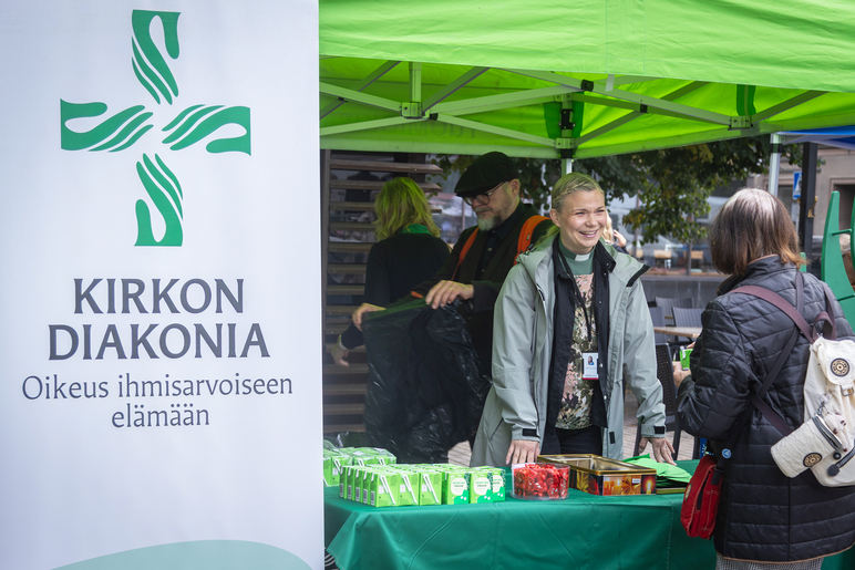 Kaksi ihmistä tiskin molemmin puolin ulkona olevassa katoksessa. Sen vieressä on juliste, jossa lukee isoilla kirjaimilla Kirkon diakonia.