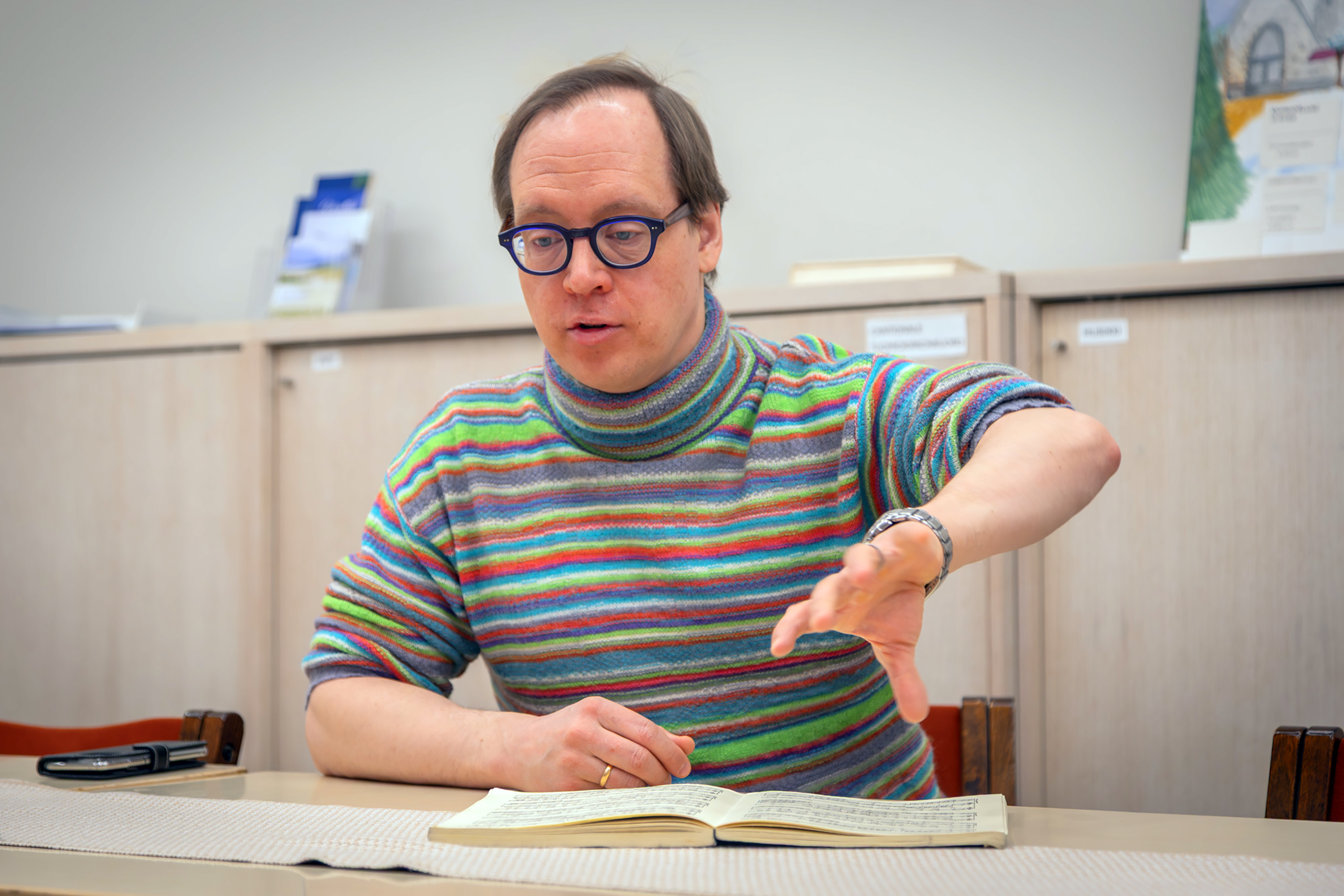 Silmälasipäinen mies istuu pöydän ääressä vihko edessään ja nostaa vasenta kättään pyörähtävällä liikkeellä.