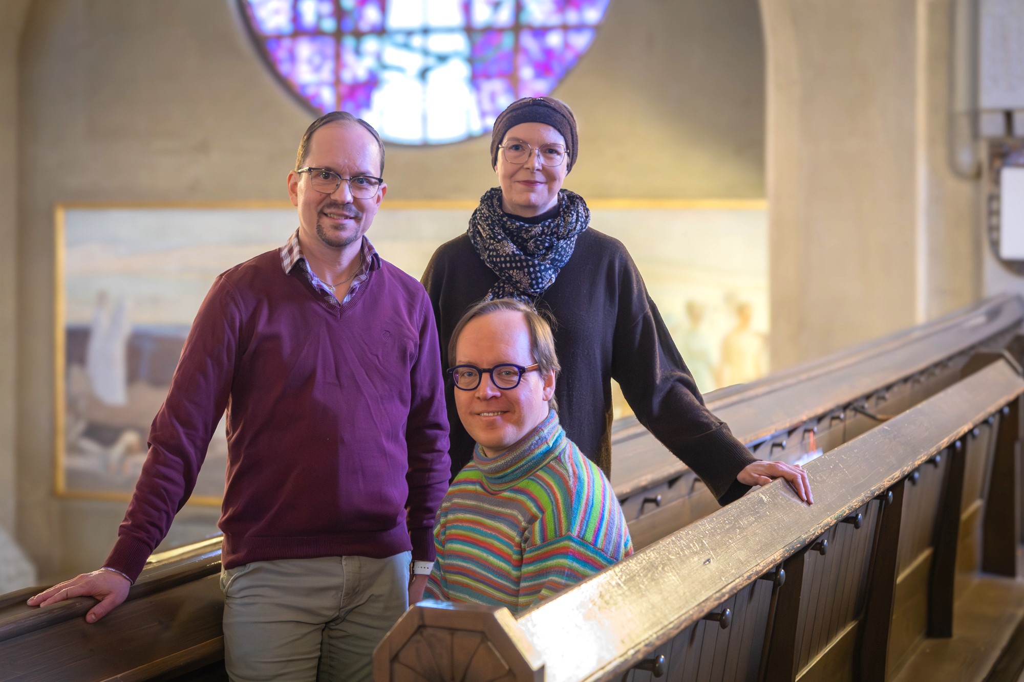 Kirkon parvella on kolme ihmistä, joista kaksi nojaa kädellään kaiteeseen ja yksi istuu. Taustalla näkyy pyöreä ikkuna.