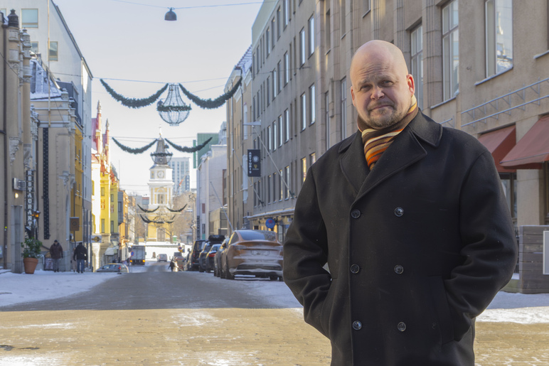 Mies seisoo katunäkymässä takki päällä kädet taskuissa. Kadun päässä näkyy puukirkko.