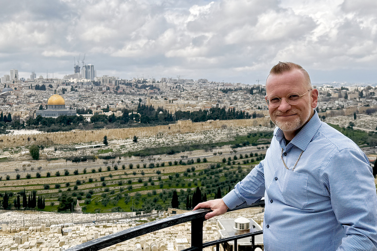 Hymyilevä mies seisoo ruutupaidassa kaupunkinäkymän edessä ja pitää kädellään edessään olevasta kaiteesta. Kauempana siintää kultakupolinen rakennus.