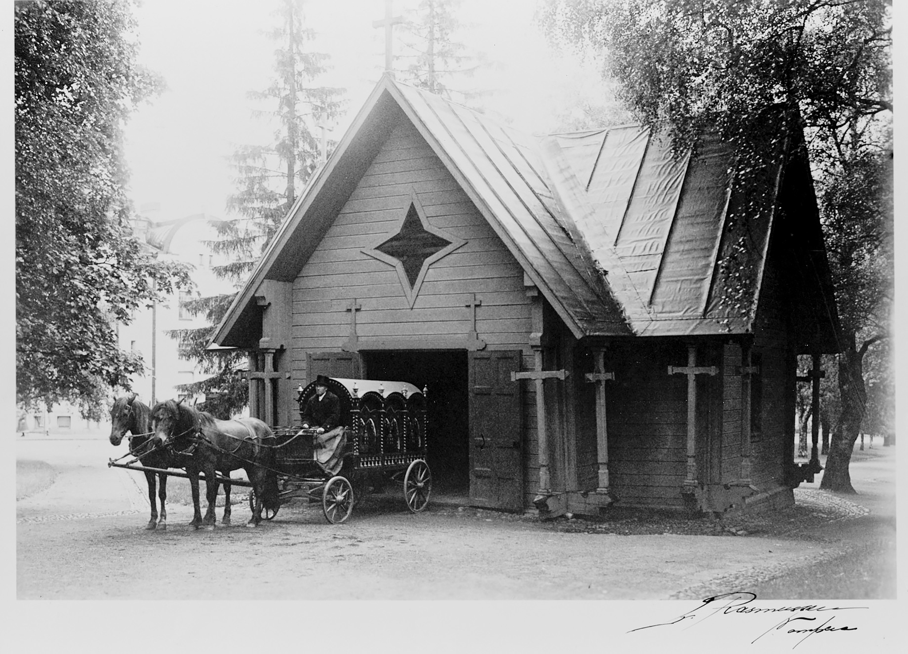 Mustavalkoinen kuva rakennuksesta, jonka jyrkkäkattoisen osan aukon edessä ovat hevosvaunut kuskeineen ja ajopiiskoineen.